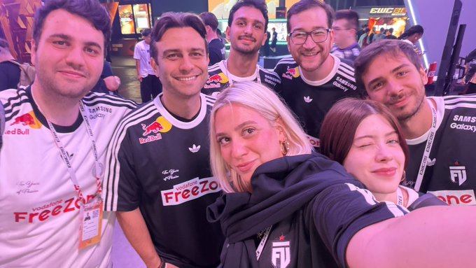 A group of seven people pose together for a selfie at what appears to be the Esports World Cup 2026 (#EWC26) event, with several wearing matching white and black jerseys featuring "Red Bull," "FreeZone," "Samsung Galaxy," and Adidas branding, along with event lanyards and badges. The cheerful group, including one woman making a playful face, appears to be a team or organization representing at the gaming tournament.