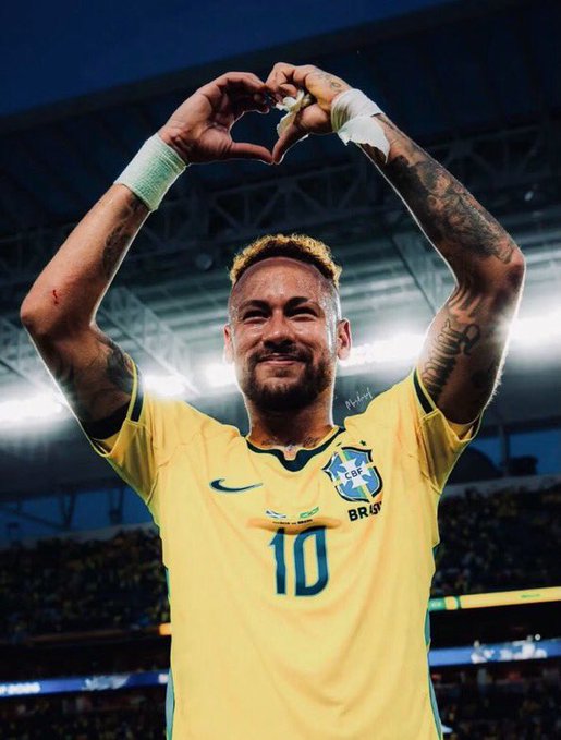 Brazilian soccer player Neymar wearing the number 10 yellow jersey, smiling and making a heart shape with his tattooed arms on a stadium field.