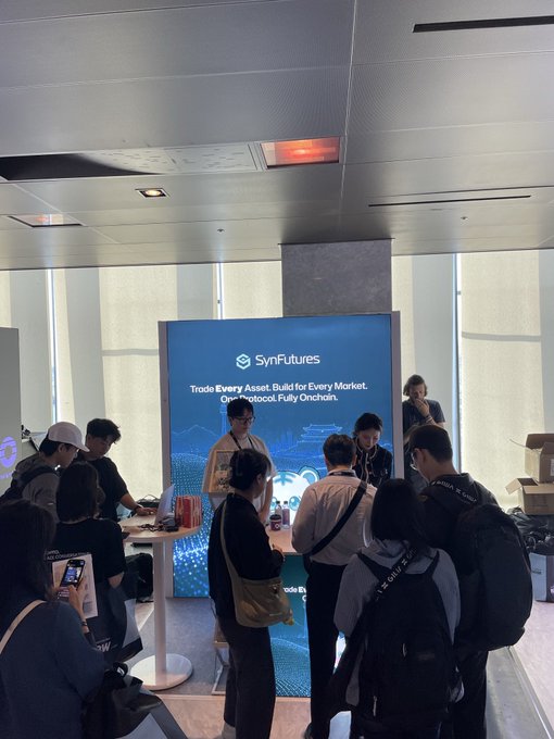 People gather around a SynFutures promotional booth displaying the text 'Trade Every Asset. Build for Every Market. One Protocol. Fully Onchain.' at what appears to be a conference or expo.