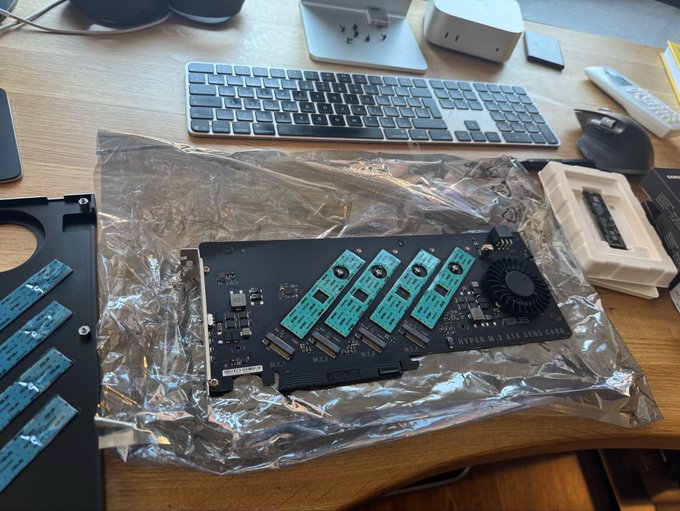 An M.2 NVMe SSD expansion card (HYPER M.2 GEN5 CARD) with four installed M.2 drives resting on an anti-static bag on a wooden desk, next to a keyboard and mouse.