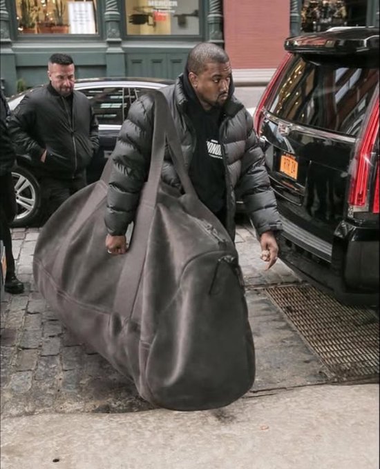 Kanye West, wearing a black puffer jacket and a mask, carries a massive oversized black duffel bag on a city street while smoking.