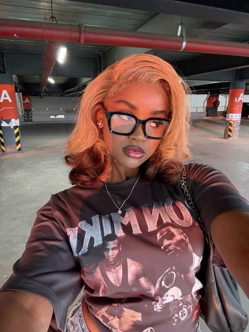 The image features a young woman wearing glasses and a t-shirt that says "River". She is posing for a photo with her left hand holding up a camera, likely taking a selfie or a picture of herself. The text in the image indicates that she has been posted on social media and asks if anyone would like to join in on the "River" campaign. It seems that the woman's outfit and the content of the post have something in common: both are related to the word "River".