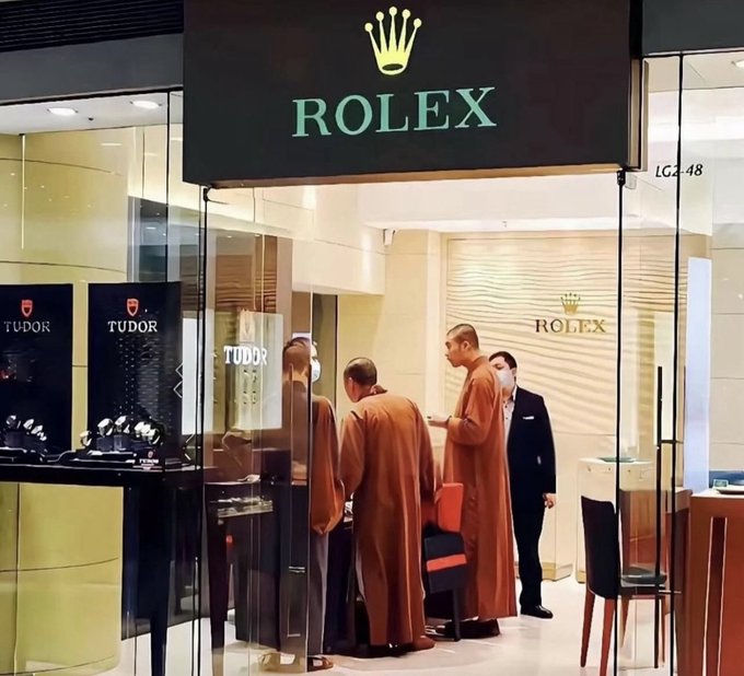 A Rolex storefront in a shopping mall with the brand's signature crown logo, featuring monks in orange robes and a staff member inside the store.