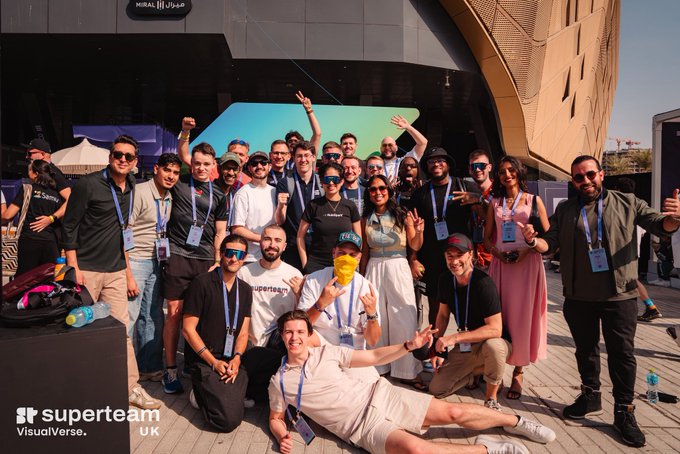 A diverse group of attendees poses energetically for a group photo at the "Superteam UK VisualVerse" event in Dubai, with several members wearing "Superteam" branded t-shirts and Solana lanyards, embodying the community-driven spirit of "finding your people" to build on the @solana ecosystem.