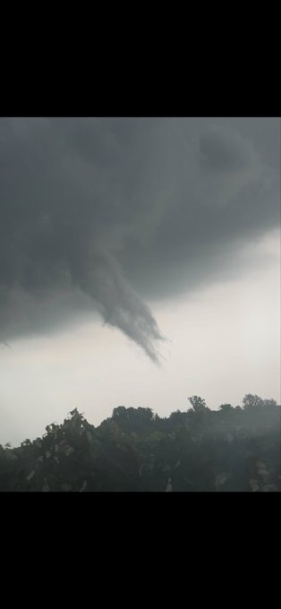 A dark, ominous storm cloud formation hanging low over a treeline, with a funnel-like shape descending from the clouds.