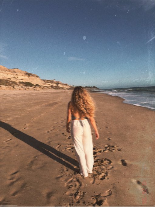 A woman with long, wavy blonde hair walks barefoot along a sandy beach, leaving footprints behind her. She wears white wide-leg pants and is photographed with a grainy, vintage film effect, with coastal cliffs visible in the background and ocean waves to the right.