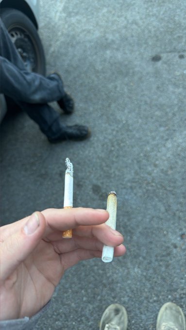 A first-person view of a hand holding two broken cigarette butts, with the smoker's own feet visible below and another person's legs and boots resting on the street nearby.