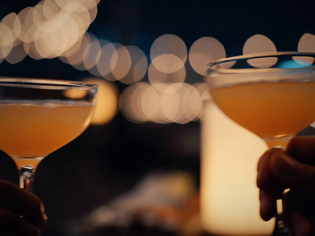 The image shows two glasses of orange liquid, possibly champagne or wine, being held by people's hands. The glasses are placed side by side with their stems pointing upwards. In the background, there is a blurred cityscape visible through the window, adding an urban touch to the scene. The caption "You can literally open a 30 day DCA on a friday night during your hinge date Just use @jup_mobile" suggests that this image might be related to a social media post or advertisement promoting a special event or occasion where people are celebrating with drinks and possibly a city tour, given the presence of the cityscape in the background.
