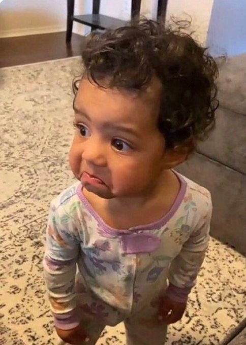 A young toddler with curly dark hair wearing patterned pajamas makes an adorable pouty face while standing on a rug in a living room.