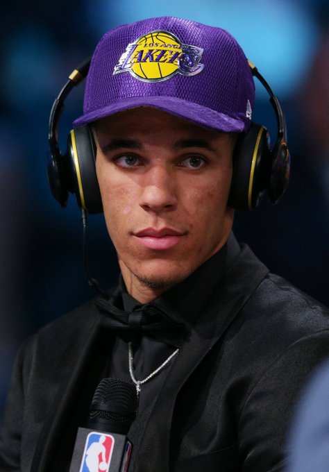 A young man wearing a purple Los Angeles Lakers cap and large black headphones, dressed in a black shirt and bow tie, speaking into an NBA microphone.