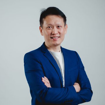 A man in a blue blazer and white t-shirt stands smiling with arms crossed against a light gray background. The image appears to be a professional portrait of Richard Teng, Binance's co-CEO referenced in the accompanying post about expanding stock access to global markets.