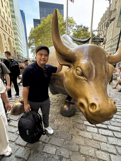 A man poses next to the iconic Charging Bull statue near Bowling Green, with an "Evergreen Pkwy" street sign visible in the background, symbolizing his visit to Wall Street's financial district as an Indonesian founder building the IDRX Rupiah stablecoin. The image conveys his ambition to tap into the heart of global finance while connecting with stablecoin and FX builders.
