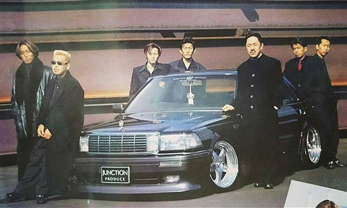 A retro-style photo of seven men in black outfits standing around a customized black sedan with a 'JUNCTION PRODUCE' license plate in what appears to be a parking garage.