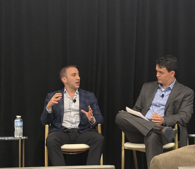 Two men in business attire sit on stage against a black backdrop, with the speaker on the left gesturing animatedly as he discusses Figure's blockchain-based capital markets platform, while the moderator on the right listens attentively with notes in hand during Goldman Sachs' Communacopia + Technology Conference.