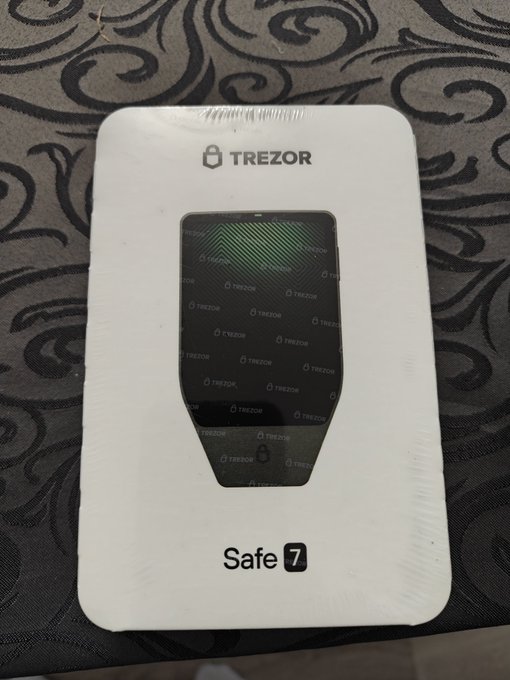 A Trezor Safe 7 hardware cryptocurrency wallet in its white retail packaging, photographed on a patterned dark surface.
