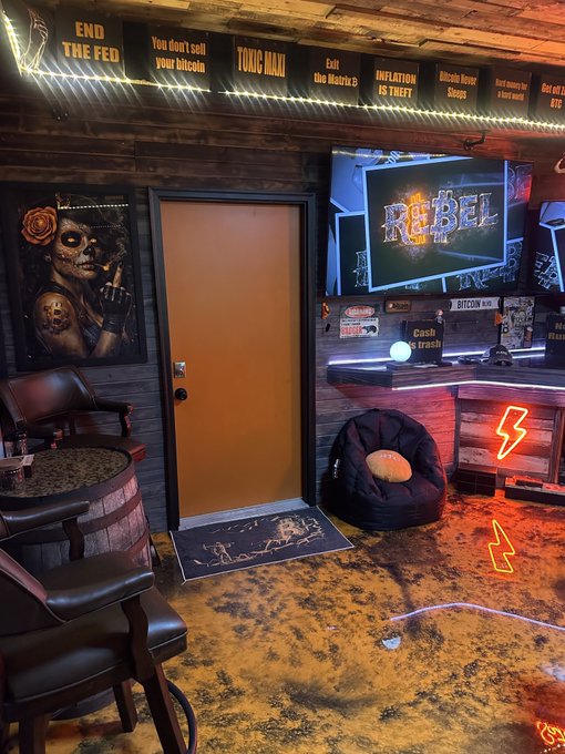 An interior room with rustic decor featuring signs reading 'END THE FED,' 'You don't sell your bitcoin,' 'TOXIC MAXI,' 'INFLATION IS THEFT,' and 'Bitcoin Never Sleeps,' alongside a screen displaying 'REBEL,' a Sugar Skull painting, and barrel furniture.