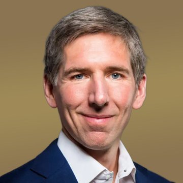 Professional headshot portrait of a smiling man with short brown hair wearing a dark blue suit and white shirt against an olive background.