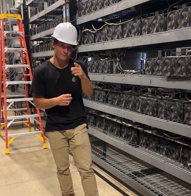 A young man wearing a white hard hat and black t-shirt giving a thumbs-up while standing in front of rows of cryptocurrency mining rigs, with an orange ladder visible on the left.