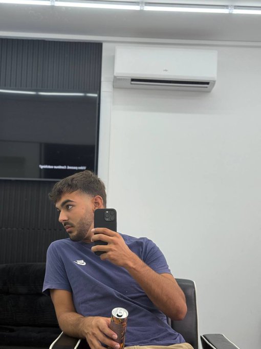 A man wearing a Nike t-shirt takes a mirror selfie while holding a can and his phone, in a room with a wall-mounted air conditioner. The casual "Good Morning, drop your $SOL address" post pairs with the laid-back, casual vibe of the morning photo, using the relaxed setting to engage followers in a friendly crypto giveaway prompt.