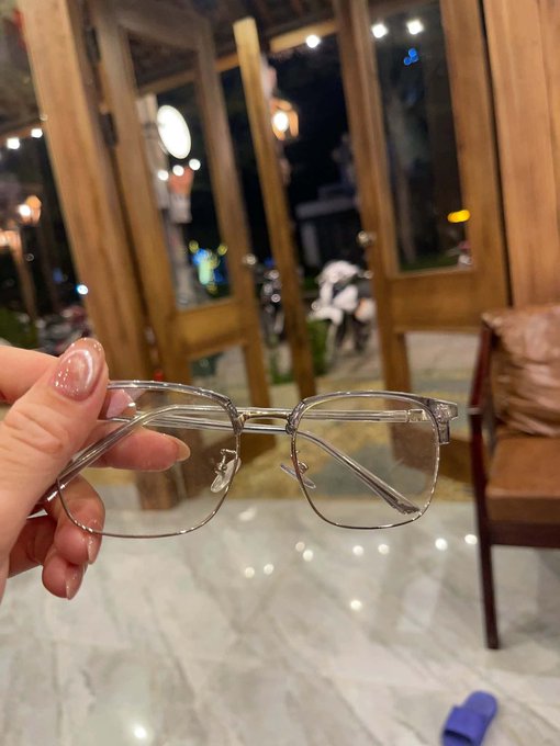 A hand with manicured nails holds up a pair of silver-framed eyeglasses, with blurred wooden doors and lights visible in the background.