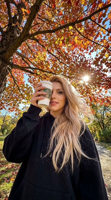 A woman with long blonde hair wearing a black hoodie holds a Starbucks coffee cup, taking a selfie outdoors under a tree with vibrant red and orange autumn leaves and sunlight filtering through.