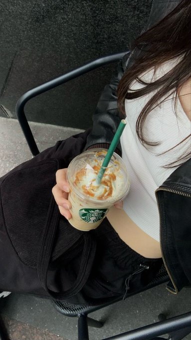 A person in a white crop top and black jacket holds a Starbucks iced caramel coffee with a green straw while sitting in a chair, capturing a relaxed Saturday morning moment that embodies the simple joys mentioned in the post—sipping coffee, unwinding, and taking a break from watching the markets.