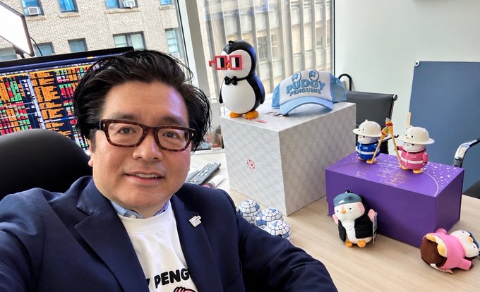 A selfie of an Asian man wearing glasses and a dark blazer over a white t-shirt with 'PENG' visible, sitting at a desk with financial market data on monitors behind him, surrounded by penguin figurines and a 'Buddy Penguin' cap.