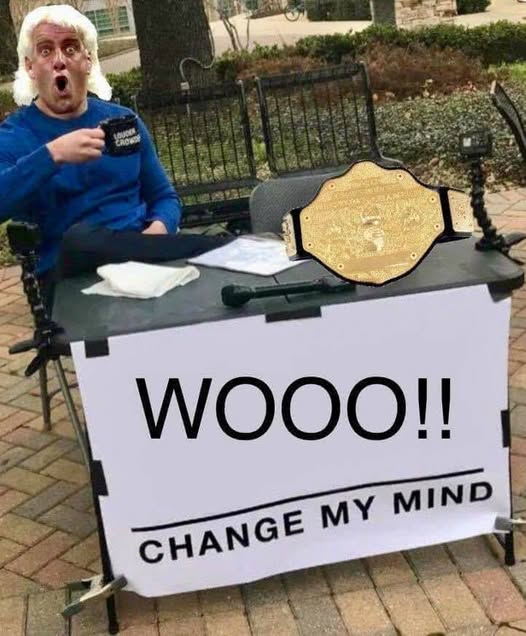 A man in a blue shirt sits at a table outdoors with a shocked expression, pointing at a sign reading 'WOOOO!! CHANGE MY MIND', with a WWE championship belt displayed on the table.