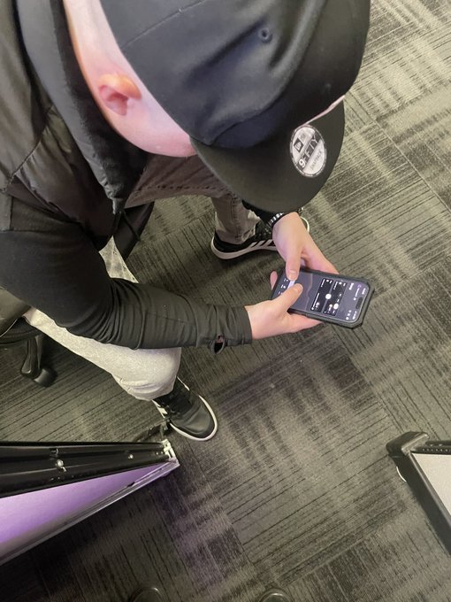 A photo taken from above showing a person in a black cap and jacket using a smartphone displaying what appears to be a trading or charts app.