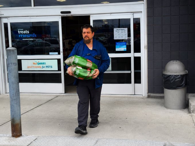 A bearded man in a blue jacket exits a PetSmart store carrying two large green bags, appearing to be catnip, while signs reading "treats rewards" and "ANYTHING for PETS" are visible in the windows behind him. The image humorously illustrates the post's premise of a man stockpiling catnip, which the joke frames as suspicious behavior warranting a Florida ASPCA advisory.