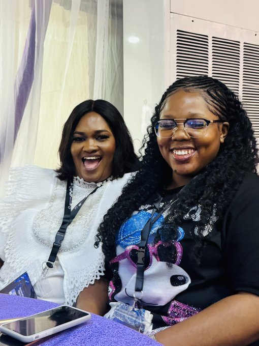 Two women are laughing joyfully together at an indoor event, one wearing a white lace top and the other a panda graphic shirt with glasses.