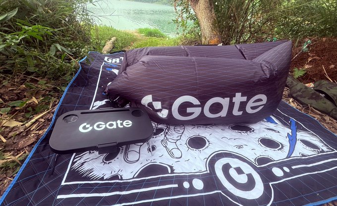 A camping setup by a turquoise lake featuring a 'Gate' branded blanket, pillow, and inflatable lounger, all black with the white Gate logo.