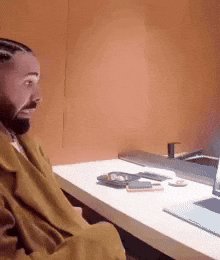 The image shows a man sitting at a desk with a laptop computer, wearing a brown coat. He appears surprised or shocked by something happening on his computer screen. The background of the image features an orange wall, which contrasts with the white table where the man is seated. There are also two cell phones placed on the table in front of him.
