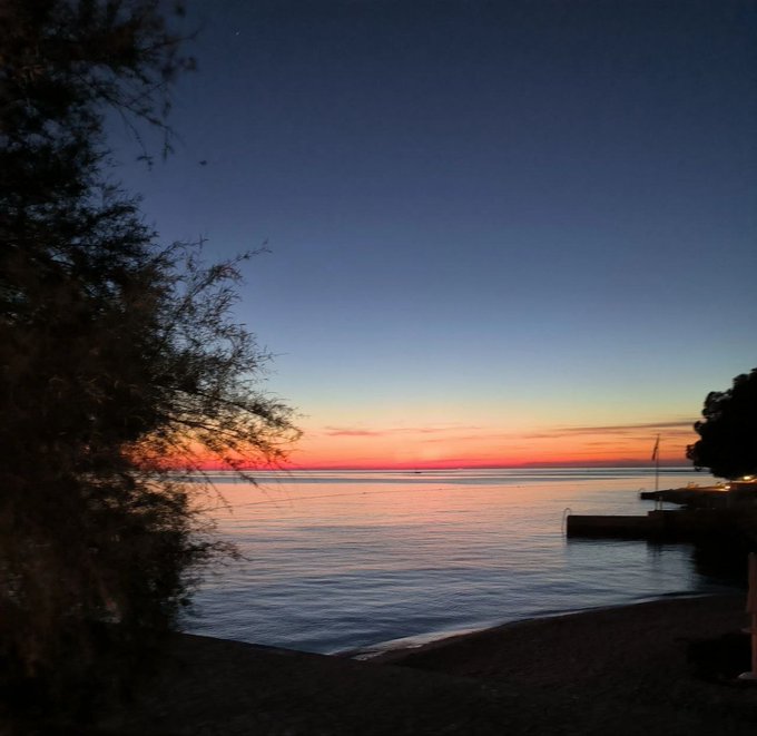 A tranquil sunset scene over a calm body of water, with silhouettes of trees framing the view and a gradient sky transitioning from deep blue to orange at the horizon.