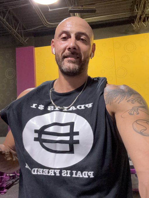 A smiling, bald man with earbuds and arm tattoos wears a black tank top featuring a coin logo with the text "PDAI IS FREEDOM" and "PDAI IS $1," directly echoing the post's message that $PDAI symbolizes both a price point and personal liberation.