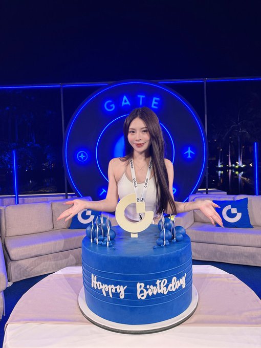 A smiling young woman with long dark hair stands behind a blue birthday cake reading 'Happy Birthday' with a 'G' topper, in a blue-lit room with 'GATE' signage on the wall.