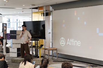 The image shows a man standing at a podium, giving a presentation or speech to an audience of people sitting behind him. The room appears to be a conference or meeting space with a large screen displaying the word "affine" written on it. There are several people in the audience, some sitting and others standing, all attentively listening to the speaker. A few chairs can also be seen around the room.