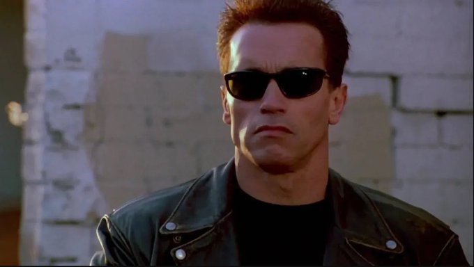 Arnold Schwarzenegger wearing a black leather jacket and dark sunglasses, standing in front of a weathered white brick wall.