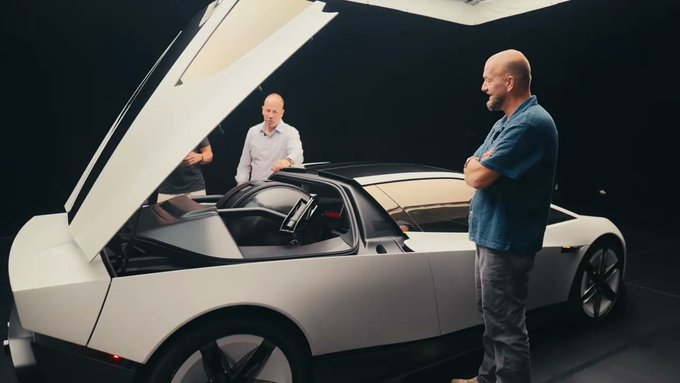 Two men stand beside a white sports car with its scissor doors open in a dark studio setting.