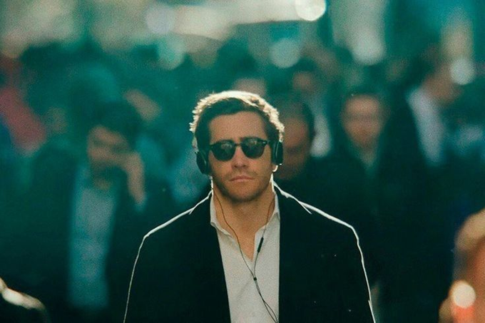 A still of a man with short hair wearing sunglasses, headphones, a white shirt, and a dark jacket, walking with a blurred crowd in the background.