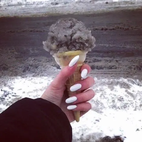A first-person view of a hand with white manicured nails holding an ice cream cone with a chunky purple scoop, set against a snowy beach background.