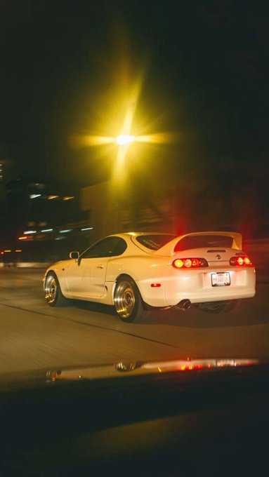 A nighttime photo of a white Toyota Supra with gold wheels driving under a glowing yellow streetlight, captured from inside another vehicle.