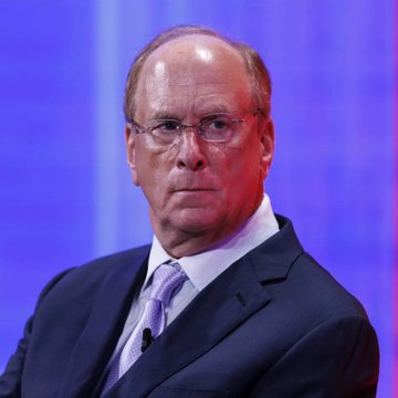 An older man with glasses, wearing a dark suit, white shirt, and light purple tie, speaks at an event with a blue and red background.