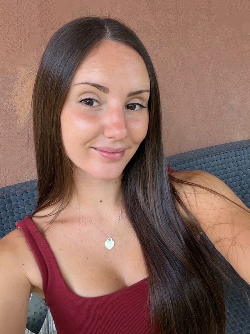 A woman with long brown hair wearing a red tank top and a silver heart necklace smiles softly at the camera, posing against a textured terracotta wall. The warm, casual selfie complements the friendly "GM to the real ones who say it back" greeting, conveying a sense of welcoming positivity to her followers.