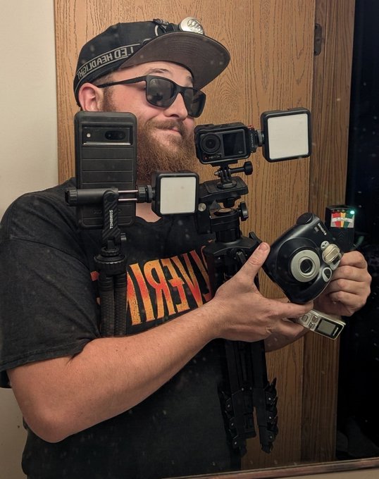 A bearded man wearing sunglasses, a cap, and a Led Zeppelin shirt poses in a mirror selfie, surrounded by an elaborate multi-camera setup including phones, GoPros, and light panels mounted on a rig.