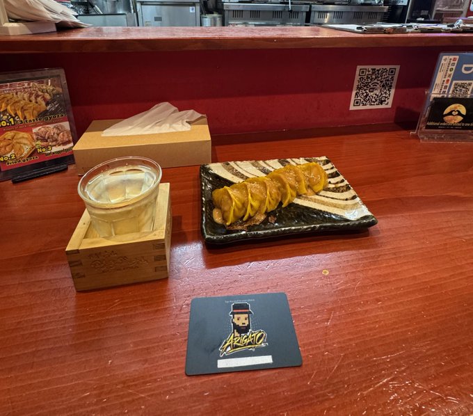 A wooden table at a restaurant displaying a black rectangular plate of sliced meat drizzled with yellow sauce, accompanied by a small cup of food in a wooden box and a black coaster with the 'ABURATTO' logo featuring a chef character.