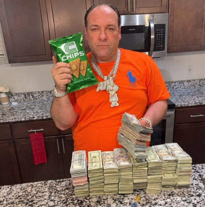 A man in an orange t-shirt wearing a large diamond-encrusted chain necklace standing in a kitchen, holding a bag of 'Chips' corn chips while displaying large stacks of bundled US currency on the counter.