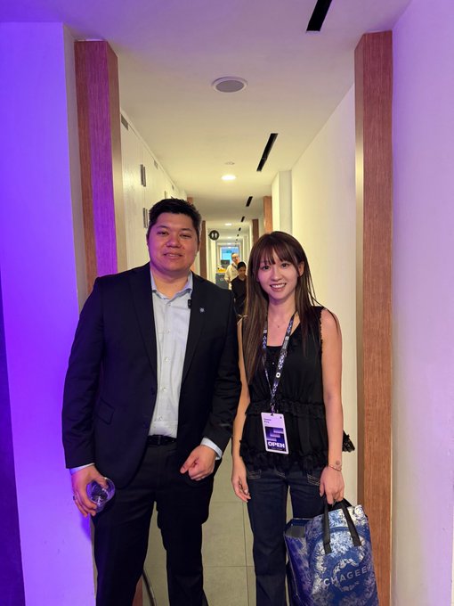 A smiling man in a dark suit and a woman wearing a lanyard with "OPEN" visible on her badge pose together in a hallway with purple-lit walls, connecting with the post about finally meeting her Monad "mama and papa" at the OPEN event.