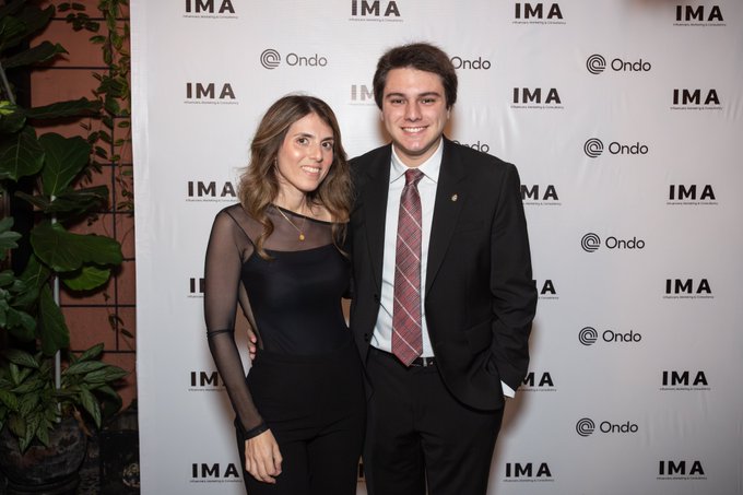A young couple poses together at what appears to be an IMA (Instituto Mauá de Tecnologia) event, standing in front of a step-and-repeat backdrop featuring IMA and Ondo logos.