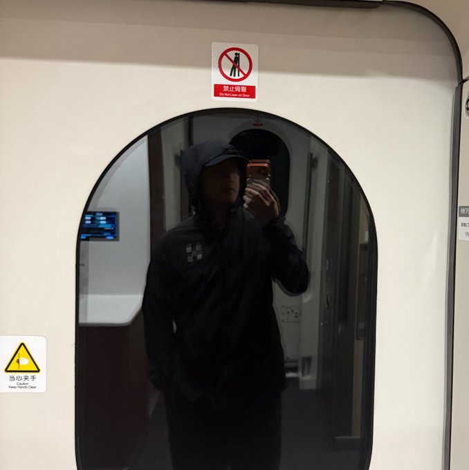 A person wearing a black hooded jacket takes a mirror selfie in a train corridor, with a 'No Lean on Door' (禁止倚靠) sign visible above.